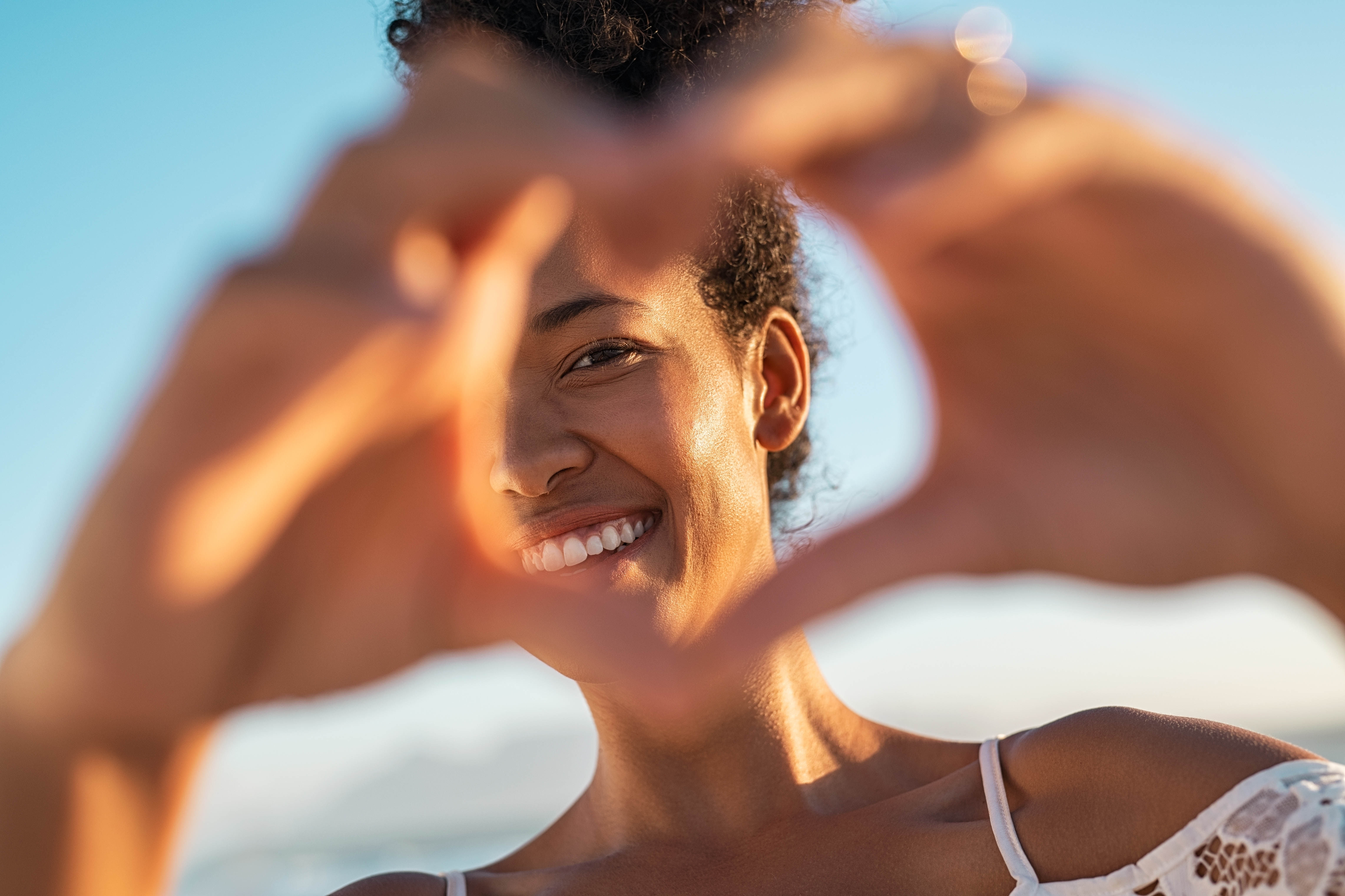 Young black woman with her hands in the shape of a heart in front of her face. Bright blue sky in the background Young black woman with her hands in the shape of a heart in front of her face. Bright blue sky in the background